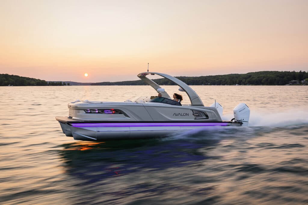 A modern speedboat named Excalibur Elite Windshield cruises swiftly across a calm lake during sunset. The boat's sleek design is highlighted by a subtle neon light strip along its side. A person is seated at the helm, steering the boat. Scenic shoreline and forested hills are in the background.