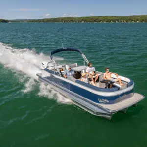 A Catalina Entertainer speeds across a lake with lush, green hills in the background. Four people are on board: one person is driving, while the others are sitting and lounging. The sky is clear, and the water is a deep blue-green.