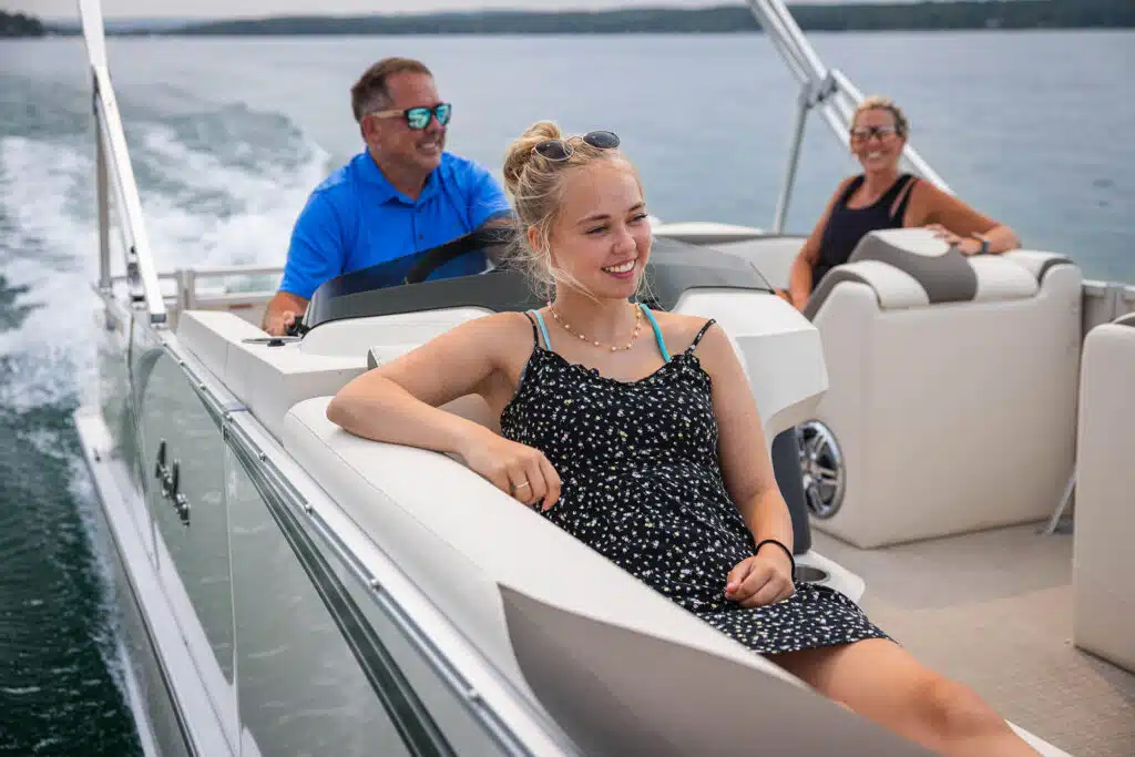 A young woman sits relaxed on the deck of an LSZ Cruise, smiling and looking ahead. A man in a blue shirt steers the LSZ Cruise, while a woman in a black top sits nearby. The LSZ Cruise is cruising on a calm body of water with a distant shore visible under a cloudy sky.