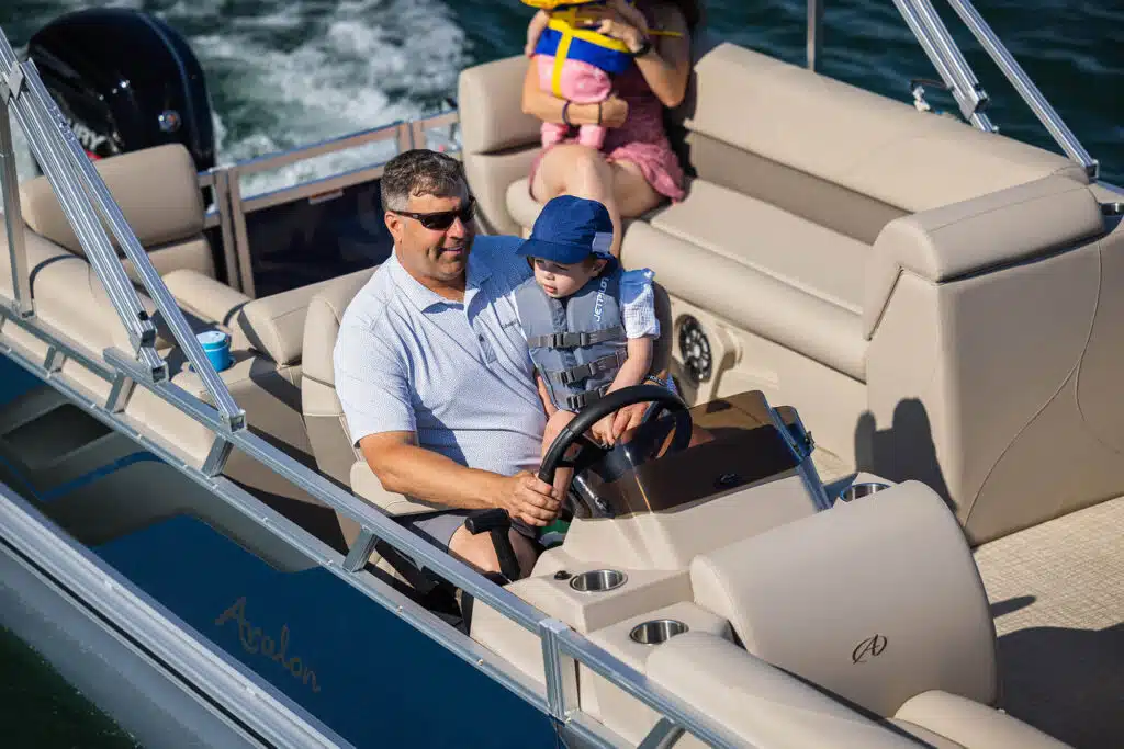 A man and a baby wearing life jackets are seated at the helm of a VLS Quad Lounge with relaxed seating. The baby is sitting on the man's lap, holding the steering wheel. Another adult, holding a child, sits in the background on a bench seat. The boat is on a body of water.