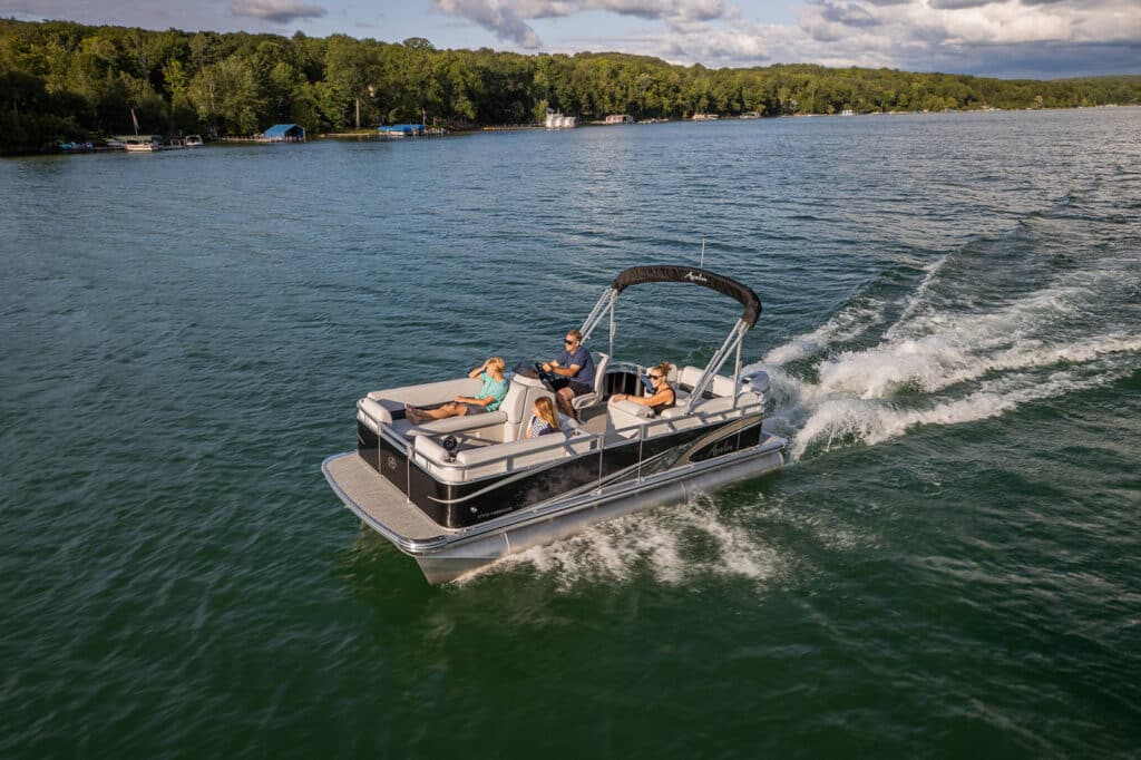 Alt description: A VTX Cruise glides across a calm lake. Two people are seated at the front, one lounging, while another person steers the boat. The lake is surrounded by a lush green forest and hills under a partly cloudy sky.

Alt title: Avalon Pontoon Boat gliding on a serene lake with people enjoying the ride.