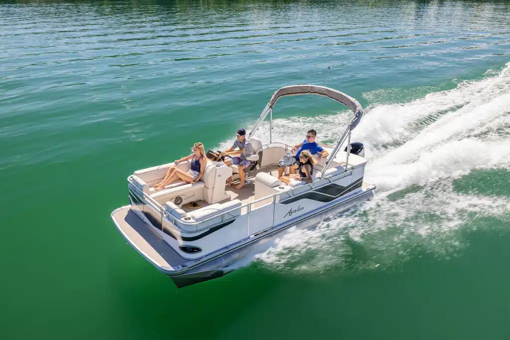 A family enjoys a sunny day on a Venture 85 Cruise gliding through calm green waters. Three people are visibly relaxed, with one steering the boat and the others lounging and enjoying the ride. A canopy provides some shade over a part of the seating area.