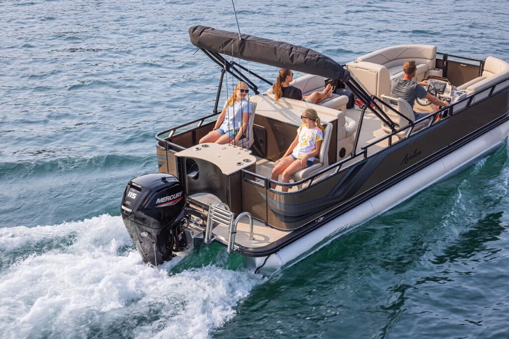 A Venture 85 Rear Fish cruising on a lake, with four people on board. Two children are seated at the back, while an adult is driving and another adult sits nearby. The boat has a black canopy, a Mercury outboard motor, and calm water surrounding it.