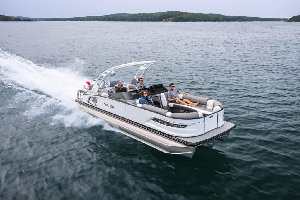 An Excalibur LTD Quad Lounge Windshield speeds across a calm lake with four people onboard. The boat has a sleek, modern design, and the passengers are seated comfortably, enjoying the ride. The background features a tree-lined shoreline under a clear sky.