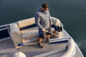 A man in a gray hoodie and black shorts uses a coiled blue hose to wash the deck of a boat. The boat features beige and navy seats, and it is on a body of water. The man is standing near the edge of the boat, focusing on cleaning the deck.