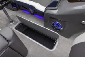 Open storage compartment in a boat with a black interior. The grey carpeted floor is partially visible, and there are blue LED lights and speaker grilles integrated into the seating and side panels. The seating and compartments are upholstered in grey and white material.