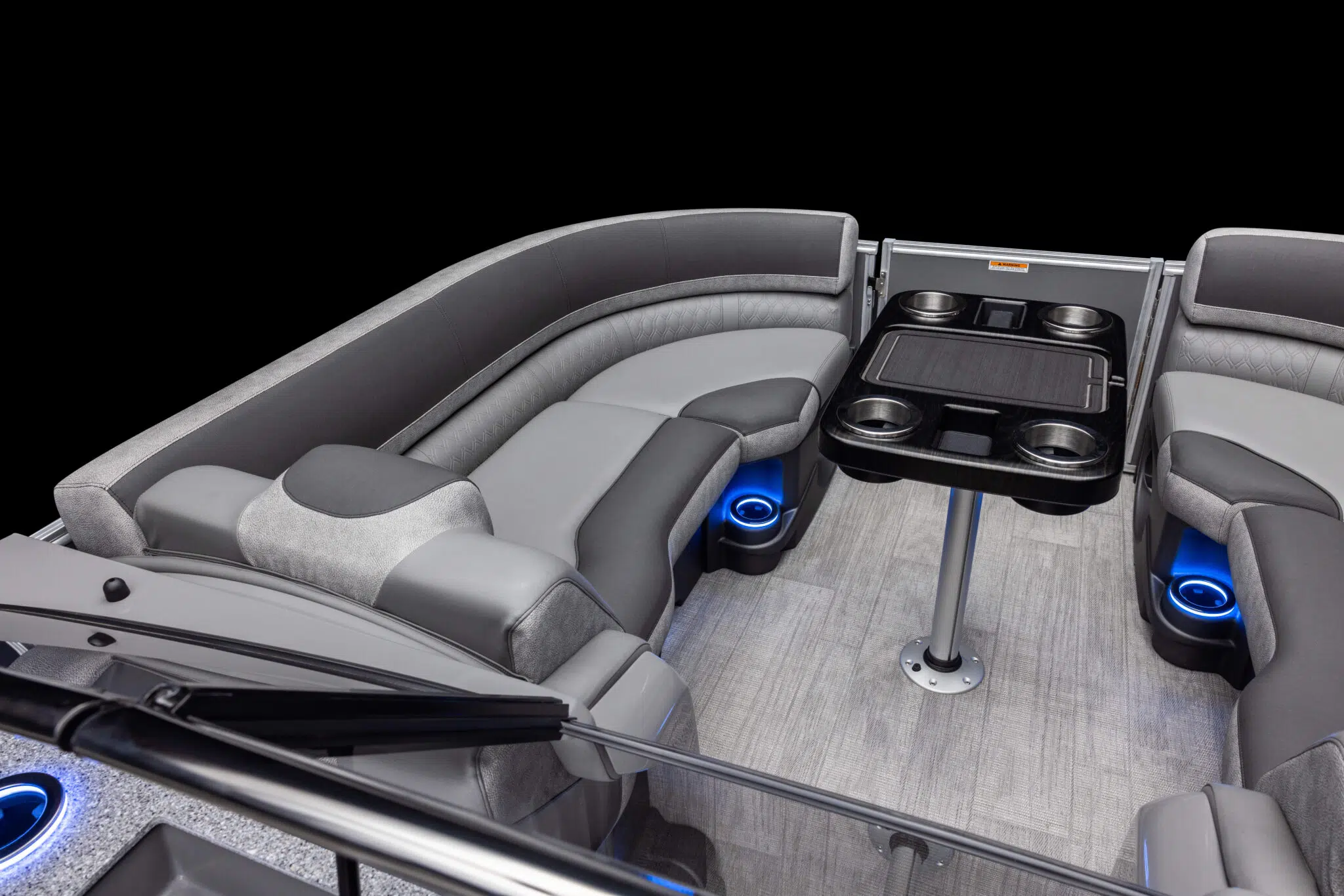 The image shows the interior seating area of a modern luxury pontoon boat, featuring curved gray and black cushioned seats. There is a central table with cup holders, and blue LED accent lighting is visible under the seats, giving the area a sleek, contemporary look.