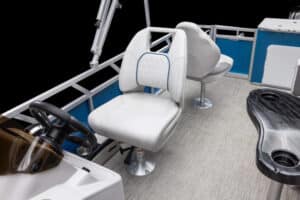 Interior view of a boat showing two cushioned white seats. One seat is positioned near the steering console with a brown windshield, and the other seat is slightly behind it. The floor has gray carpeting, and there are blue and silver railings around the perimeter.