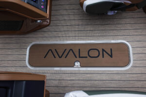 View of a boat interior showing a rectangular compartment hatch on the floor. The hatch has a wooden finish with the word "AVALON" printed in large letters. Surrounding the hatch is a textured flooring surface with horizontal lines.