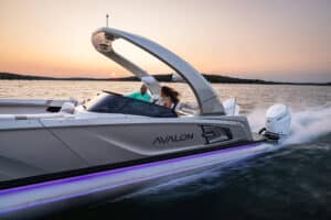 A sleek, white motorboat labeled "Avalon" speeds across a body of water at sunset. Two people are seated on the boat, enjoying the ride. The sun is low on the horizon, casting a warm glow across the water and sky. The boat's trim features a subtle purple light.