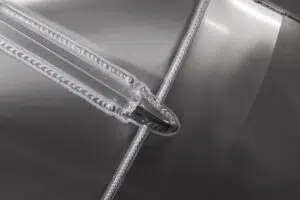 Close-up of a precise metal weld with clean, consistent seams and silver-gray coloring, highlighting the craftsmanship in industrial metalwork. The weld intersects at a right angle, demonstrating detailed workmanship.