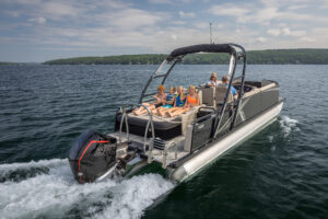 A group of people is relaxing on a pontoon boat with a canopy, cruising on a lake. The boat features a powerful outboard motor, and the passengers are enjoying the sunny weather, surrounded by lush green trees in the background.