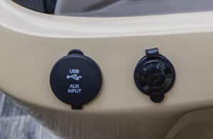 Close-up of a vehicle dashboard featuring two circular ports. The left port is labeled "USB AUX INPUT," and the right port is labeled "12V DC." Both ports have protective covers. The dashboard is beige with a black trim.