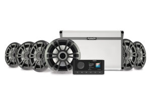 The image shows a Fusion marine audio system comprising a white amplifier, six grey and black speakers, and a black control unit with a small screen displaying various settings and information. The equipment is arranged neatly against a white background.