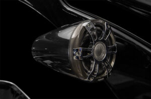 Close-up of a modern, black and chrome car speaker mounted on a shiny black surface. The design features a sleek, metallic grille with a central mesh covering. The dark, reflective background emphasizes the speaker's stylish and futuristic appearance.