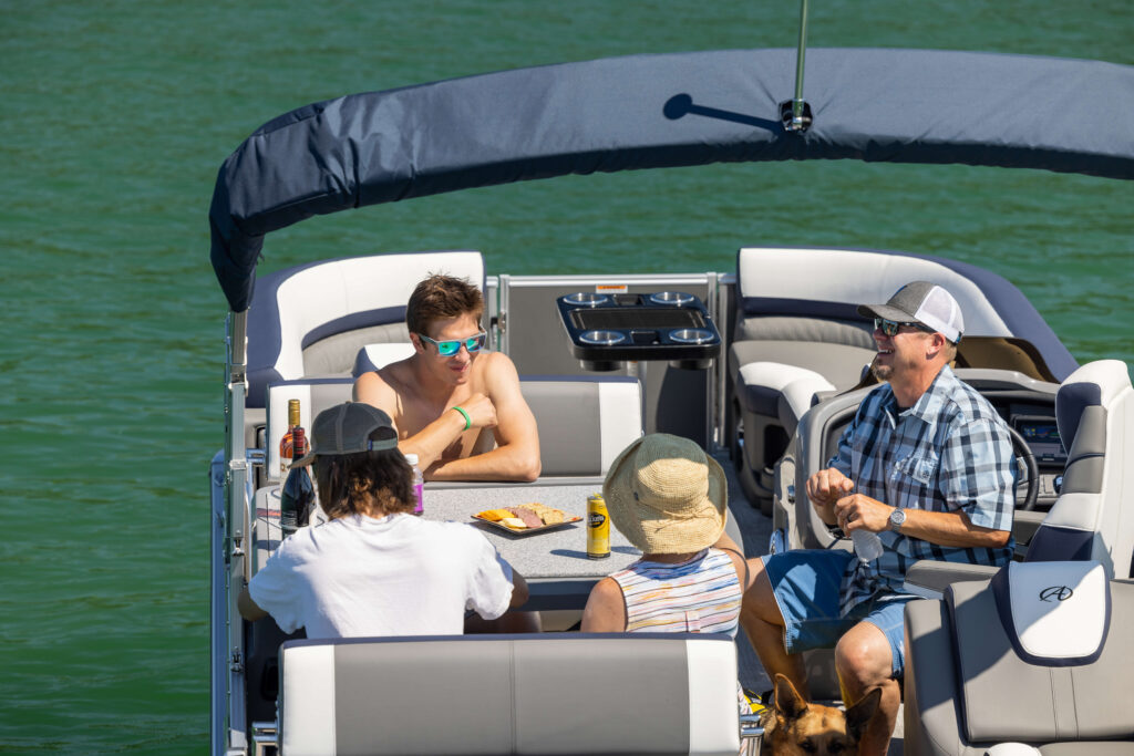 Four people are sitting and chatting on a boat. They are enjoying snacks and drinks on a table in front of them. The boat is on calm, green water. A dog is partially visible near their feet. The atmosphere is relaxed and sunny.