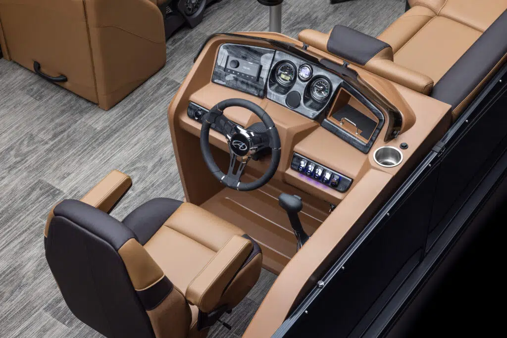 A view of a boat's cockpit featuring a tan and black captain's chair, a steering wheel, and a control panel with multiple gauges and switches. The cockpit includes a cup holder and is surrounded by tan upholstery. The boat's flooring is gray textured material.