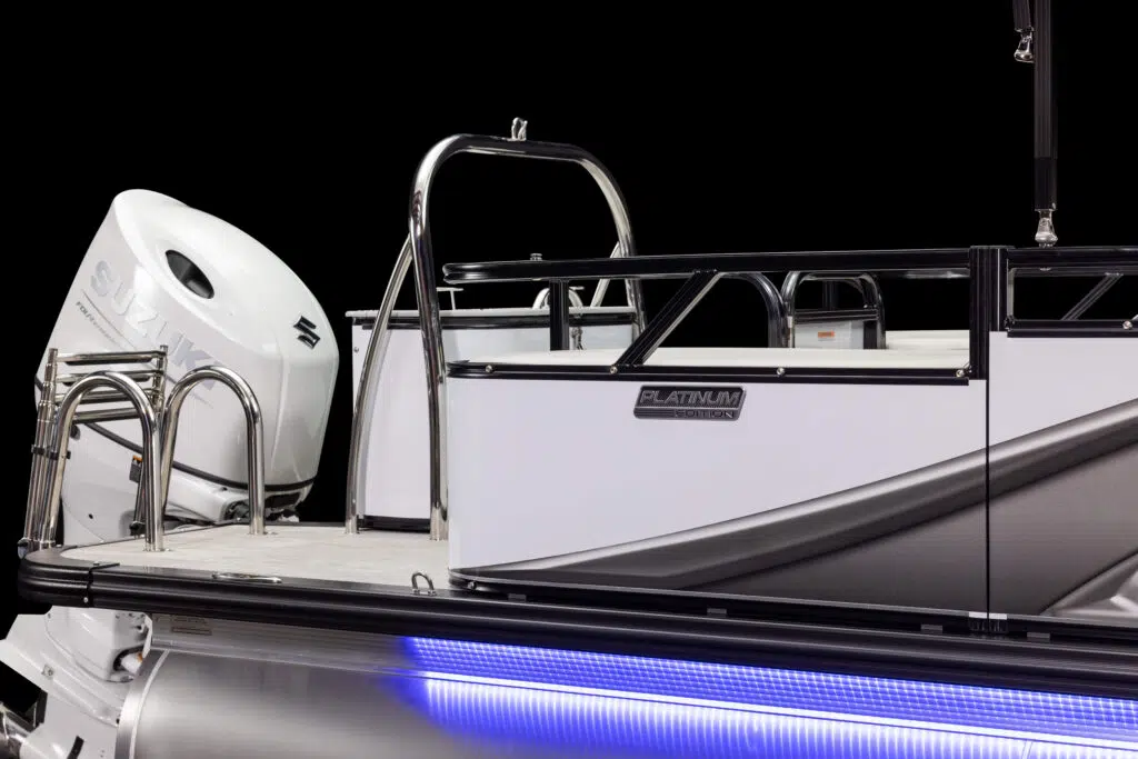 A close-up view of a modern pontoon boat. Visible features include a shiny metal railing, a "Suzuki" outboard motor, and sleek body panels with a "Platinum" label. The boat is illuminated with blue LED lights along its side, set against a dark background.