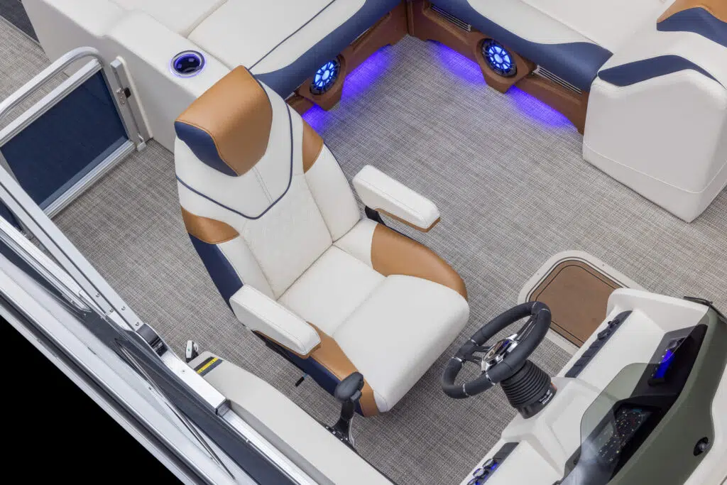 A captain's chair with white and brown cushioning is situated in a modern boat cockpit. The area features beige carpeting, ambient blue lighting, and a sleek control panel with a steering wheel. Additional seating with matching upholstery is visible around the sides.