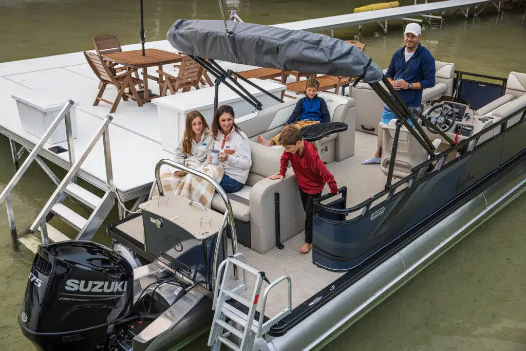 A group of five people, including two adults and three children, are on a Venture Cruise Rear Bench docked next to a pier. The adults are sitting and holding mugs, while the children are positioned around them, one climbing down the back. The boat is equipped with a Suzuki motor.