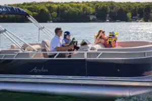 A family of four is enjoying a sunny day on a lake in a pontoon boat. The boat, branded "Avalon," is cruising on calm waters. One adult is steering, another is holding a small child wearing a life jacket, and a child in a hat is seated nearby. Trees line the distant shore.