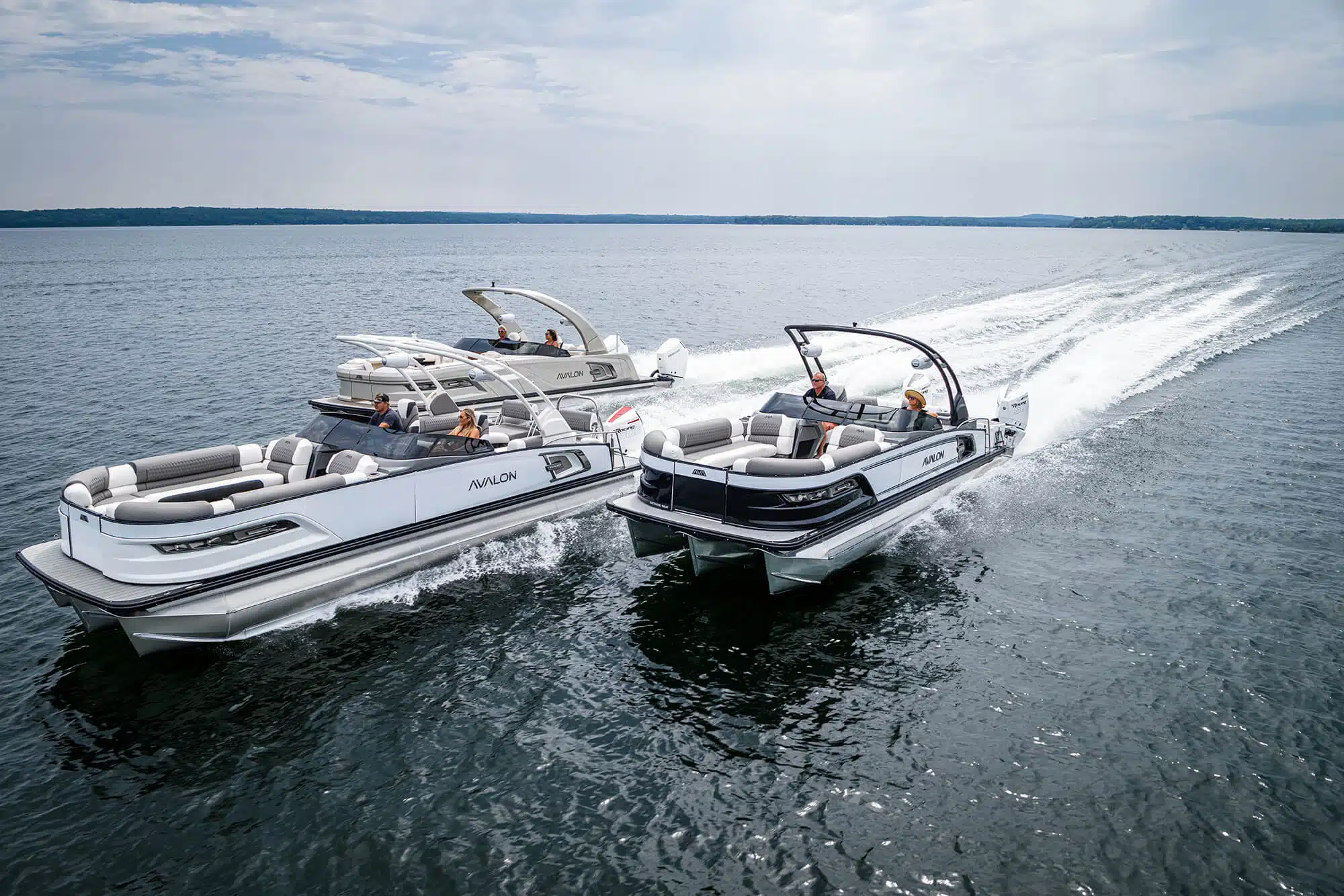 Two luxury pontoon boats, one white and one black, speed side-by-side across a large body of water. Both pontoons near me carry passengers, and one has an extended canopy. The sky is partly cloudy, and the distant shoreline is faintly visible.