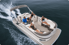 A boat with six people onboard is cruising through the water. Some are sitting on the cushioned seats while others are leaning back, enjoying the ride under the shade canopy. The sturdy pontoons provide stability as they glide over calm waters that perfectly reflect the blue sky.