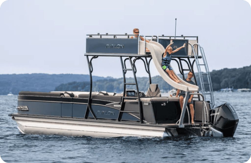 A double-decker pontoon boat is anchored on a lake. Three people are on the boat: one at the top deck, one sliding down a water slide into the lake, and one standing near the slide. The pontoon has "Avalon" written on it and boasts a scenic background of tree-covered hills.