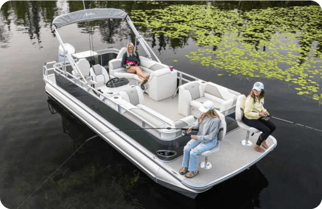 Three people are on a pontoon boat on a calm water body. Two individuals are seated at the rear of the pontoons with fishing rods, and the third person is seated towards the middle of the boat, relaxing. The surrounding water has patches of aquatic plants.