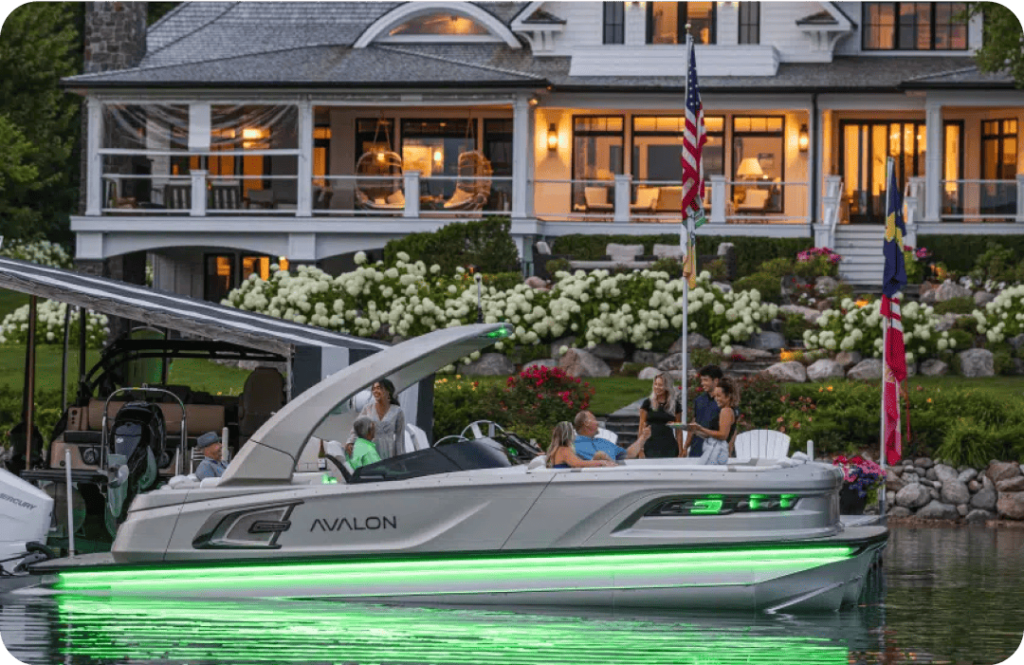 A sleek motorboat with green underglow lighting is docked by a large, well-lit lakeside house. People are sitting and standing on the boat's pontoons, enjoying an evening gathering. The house features a spacious porch and well-maintained garden. An American flag is in view.