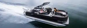 A sleek black pontoon boat speeds across the water with two people aboard. One person stands at the helm steering while the other lounges on a cushioned seat, relaxing under the sun. The sturdy pontoons create a trail of white waves behind it, cutting through the calm water.