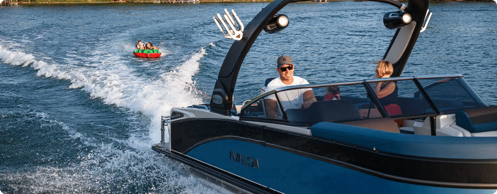 A person wearing a cap drives a motorboat with blue and black accents on a body of water, while another person sits beside them, surrounded by sleek pontoon accessories. In the background, three people are riding an inflated tube being towed by the boat, creating a splash trail behind them.