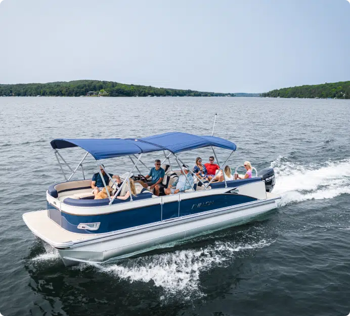 A group of people are sitting and relaxing on one of the best pontoon boats, featuring a blue and white design with a canopy. They're cruising on a large body of water surrounded by forested shoreline in the distance under a clear sky.