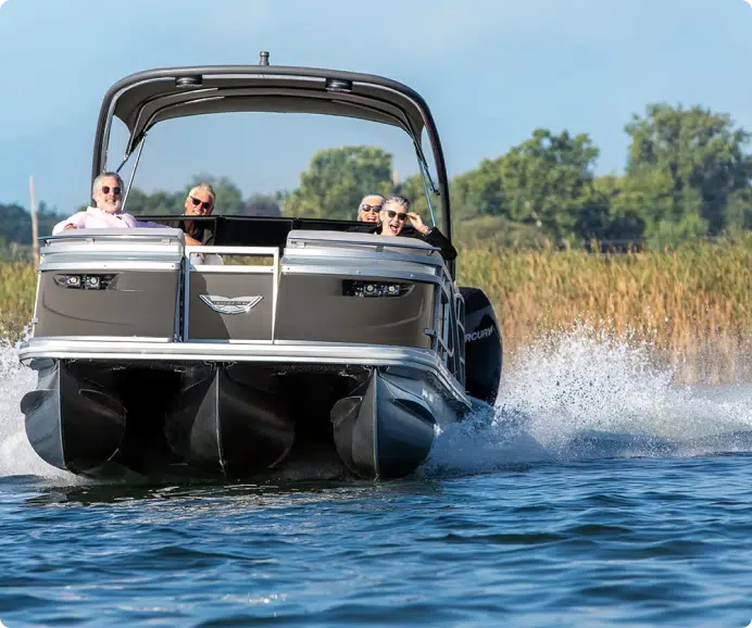 A group of six people enjoy a ride on one of the best pontoon boats, creating a splash in the water. The motorized boat glides on a calm body of water on a sunny day, with greenery and trees visible in the background.