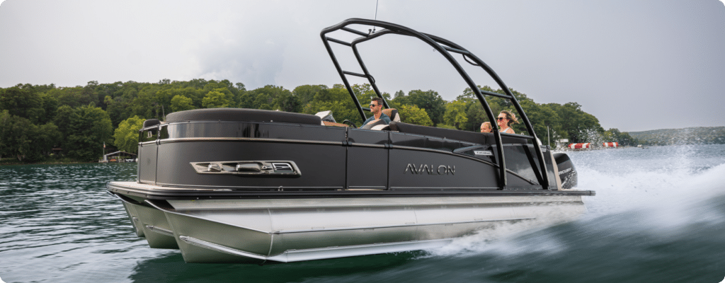 A sleek black pontoon boat, considered one of the best pontoon boats, glides swiftly across a lake with three people on board, surrounded by lush green trees and clear water under a cloudy sky. The boat's wake leaves a trail behind it, creating a sense of speed and excitement.