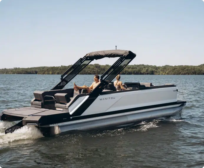 A sleek, modern pontoon boat glides over the water with two passengers relaxing under a canopy. The Manitou, one of the best pontoon boats on the market, boasts a streamlined design and is surrounded by calm waters and a distant treeline.