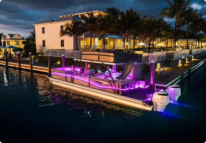 A luxury boat, one of the best pontoon boats around, illuminated with vibrant pink and blue lights, is docked by a waterfront property in the evening. The waterfront area is lined with palm trees and upscale buildings, creating a serene and elegant atmosphere.