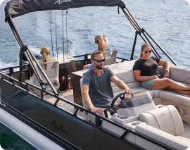 A family is enjoying a ride on a boat. A man with a beard is steering the boat, while a woman wearing sunglasses is seated next to him. Two children are sitting on the deck at the back of the boat. Fishing rods are also visible in the background. This scene could be straight out of an ad for pontoons near me.