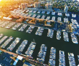 An aerial view of a bustling marina filled with numerous boats and yachts docked along pontoons near me. Surrounding the marina are buildings and trees, with the sun setting in the background, casting a warm glow over the scene.