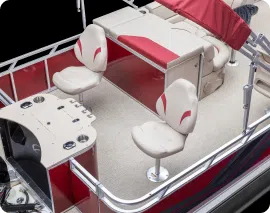 A pontoon boat interior featuring two cream-colored captain's chairs with red accents, a steering console with controls, a table with cushioned red and cream benches, and part of a red canopy overhead. The sleek red and white color scheme is perfect for those seeking stylish pontoons near me.