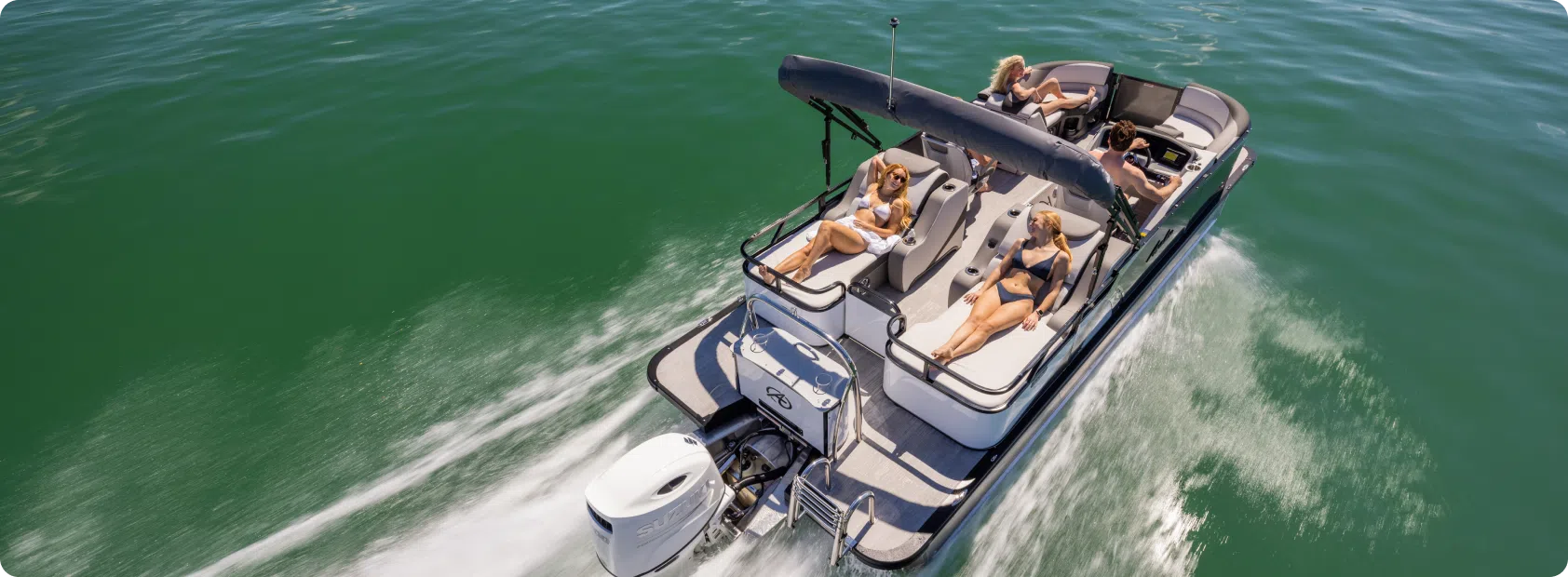 Aerial view of a motorboat cruising on clear green waters. The boat, resembling luxurious fishing pontoons, has a canopy and several passengers relaxing on deck chairs and in the lounge area. Two women sunbathe on the front deck while others sit and enjoy the ride. The boat's wake can be seen behind.