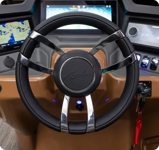 Close-up of a boat's steering wheel featuring a sleek, modern design with a metallic and black finish. The dashboard displays digital screens showing navigation maps and controls, with various buttons and switches. A key with a red lanyard hangs to the side, perfect for any discerning pontoon dealer.