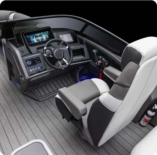 The image shows the interior of a luxury boat cockpit with modern navigation and control equipment, likely offered by a seasoned pontoons dealer. There is a comfortable, cushioned captain's chair, a digital display screen, and various buttons and levers for operating the boat. The flooring is sleek and gray.