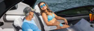 A woman wearing a blue bikini and reflective sunglasses lounges on a boat seat, smiling, while a man in a cap, sunglasses, and light blue shirt looks at her. They are enjoying a sunny day on the water, discussing their recent research on pontoon boat prices.