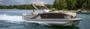 A pontoon boat with sleek aluminum pontoons cruises on a lake surrounded by a forest. The boat has a canopy and is carrying three people: a young girl in the front, a man steering, and another man sitting beside him. An American flag is attached to the boat's canopy.