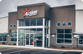 A modern commercial building with a prominent sign reading "Action" featuring a red star. The structure boasts large glass windows and doors, circular windows on the upper side, and a parking lot in front where you might find an ad for a pontoon boat for sale. The sky is overcast.