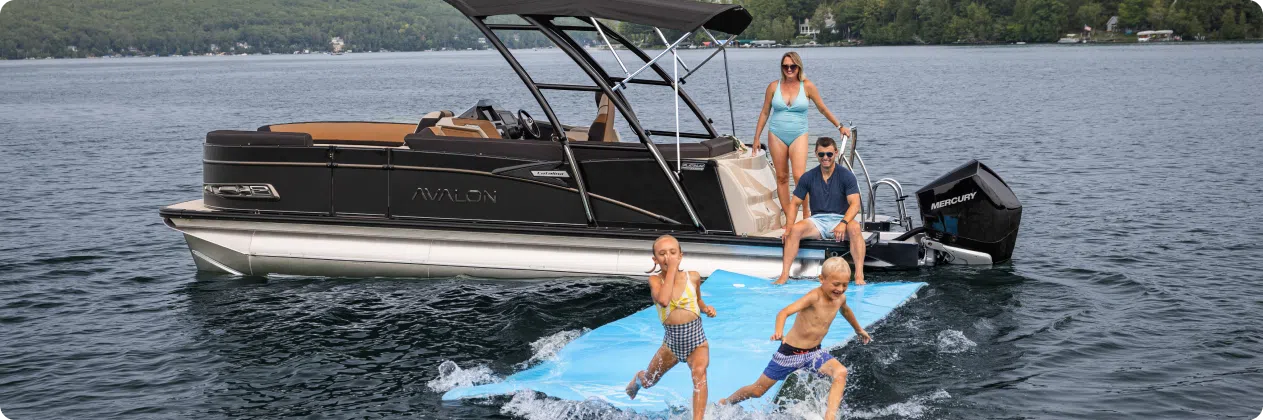 A family enjoys a day on a pontoon boat in a lake, surrounded by lush green hills and trees. Two children run on a blue floating mat in the water. A woman in a swimsuit stands on the boat while a man, relaxed near the edge with some handy pontoon accessories, watches the children play gleefully.