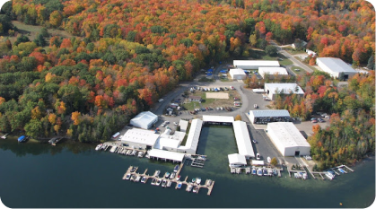 Aerial view of a lakeside marina with multiple white-roofed buildings and boat docks extending into the water, featuring a pontoon boat for sale. The surrounding area is covered in dense forest with autumn foliage in vibrant shades of red, orange, yellow, and green.