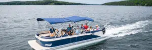 A group of people enjoy a day on a blue and white pontoon boat on a lake surrounded by forested shoreline. With the pontoon boat price being affordable, the vessel features a blue canopy providing shade. The individuals are seated and standing, some under the canopy and others at the front and back.