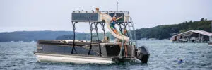 A family enjoys a pontoon boat ride on a lake. Two children play on the slide attached to the top deck of the boat, with one child mid-slide and another waiting. An adult supervises from the deck, which is supported by sturdy aluminum pontoons. Around them are green hills and a dock with a covered boat.